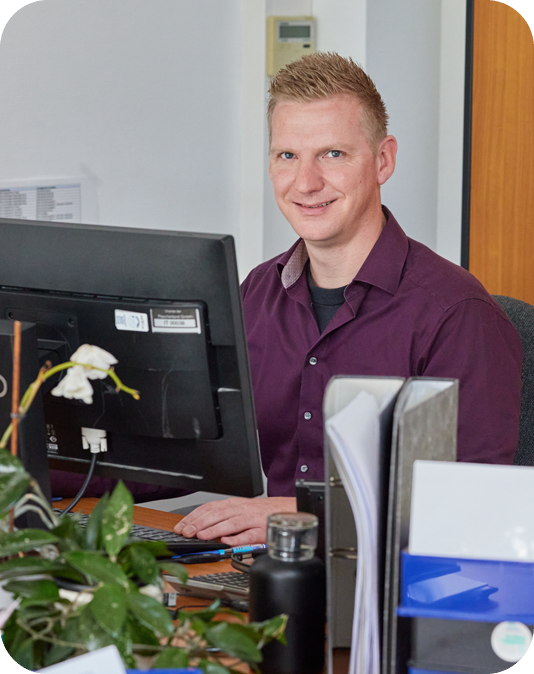 Team Leader Customer Service Friendly man with aubergine-colored shirt sitting at the computer and working
